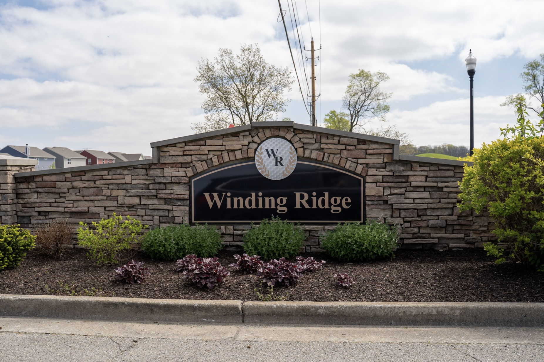 Winding Ridge entrance sign amid clouds, surrounded by neat shrubs, conveys tranquility, welcome, and a sense of home.
