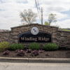 Winding Ridge entrance sign amid clouds, surrounded by neat shrubs, conveys tranquility, welcome, and a sense of home.
