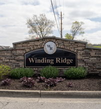 Winding Ridge entrance sign amid clouds, surrounded by neat shrubs, conveys tranquility, welcome, and a sense of home.