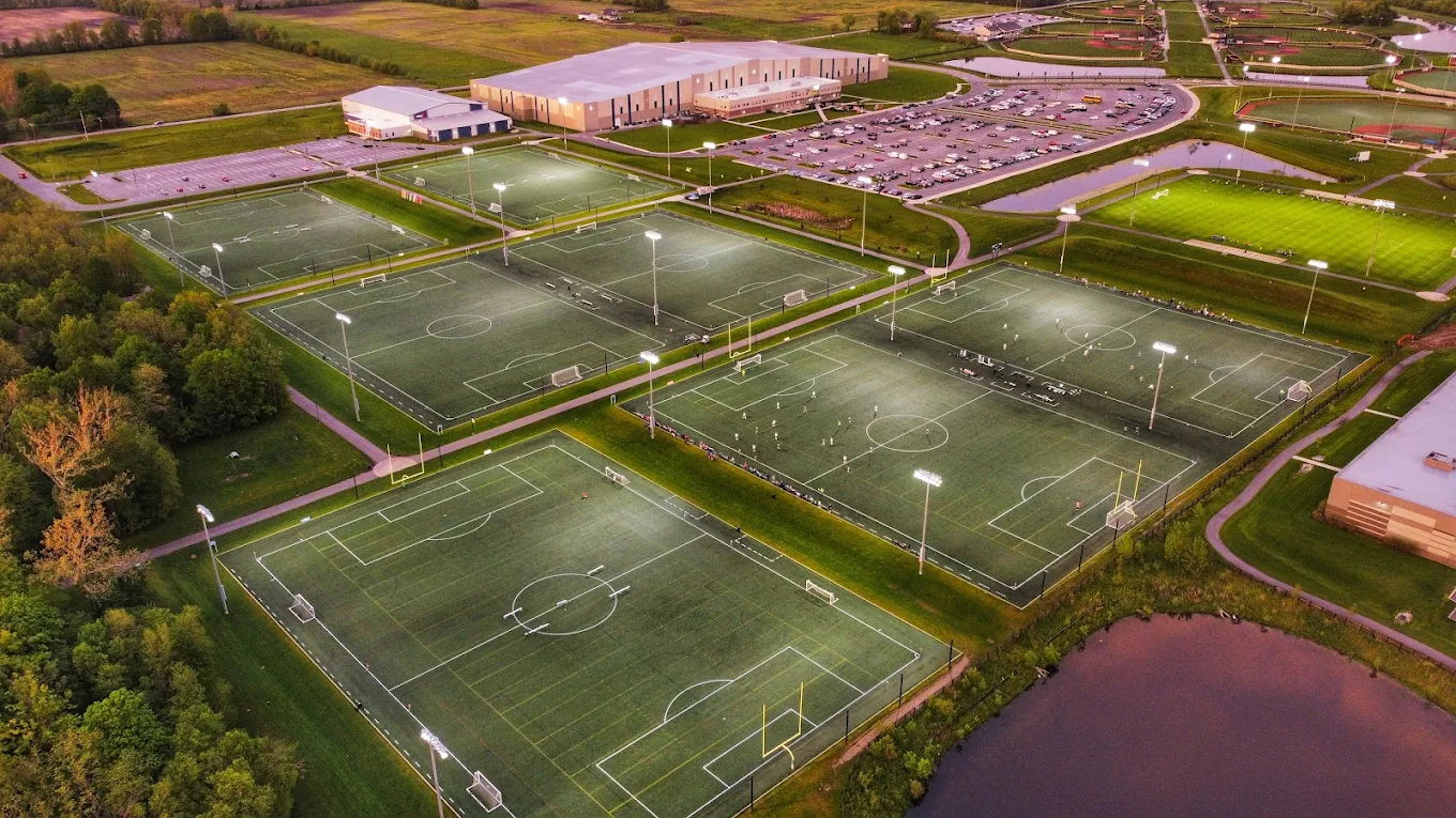 Panoramic view of a vibrant sports complex with illuminated soccer fields, greenery, and water, evoking energy and community.