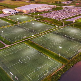 Panoramic view of a vibrant sports complex with illuminated soccer fields, greenery, and water, evoking energy and community.