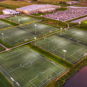 Panoramic view of a vibrant sports complex with illuminated soccer fields, greenery, and water, evoking energy and community.