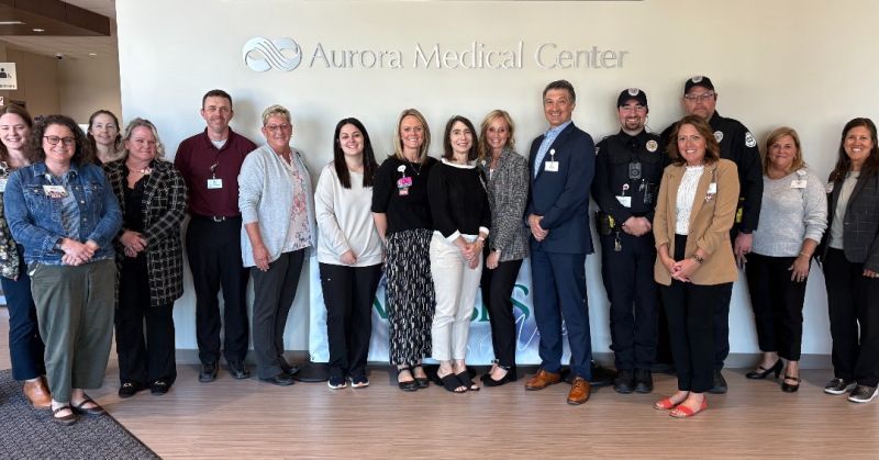 A diverse group in professional attire smiles together at Aurora Medical Center, suggesting teamwork and collaboration.