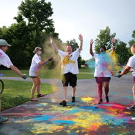 A colorful celebration scene with people throwing powders, dressed in white, expressing joy in a green outdoor setting.
