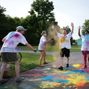 A colorful celebration scene with people throwing powders, dressed in white, expressing joy in a green outdoor setting.