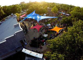 Vibrant outdoor festival with colorful canopies, a large crowd, and lush trees creating a joyful and lively community atmosphere.