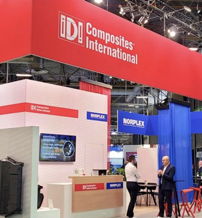Trade show booth for "ID! Composites International" with a red sign, modern display, and professional networking atmosphere.