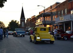 Nostalgic scene of a small-town vintage car show with classic cars, people socializing, and charming brick buildings.