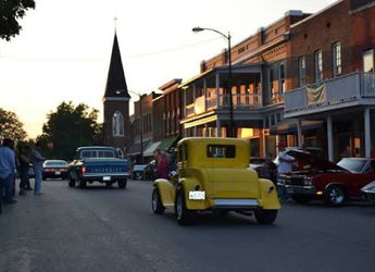 Nostalgic scene of a small-town vintage car show with classic cars, people socializing, and charming brick buildings.