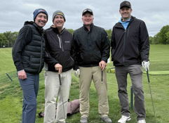 Four friends in golf attire smile on a course, exuding camaraderie and joy amidst a cloudy outdoor setting.