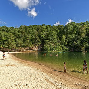 A serene beach scene with lush greenery, people relaxing on the sand, wading in water, under a clear sky with fluffy clouds.