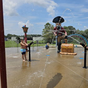 Two children joyfully play under water jets at a colorful pirate-themed splash pad on a sunny day.