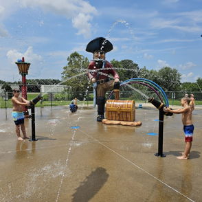Two children joyfully play under water jets at a colorful pirate-themed splash pad on a sunny day.