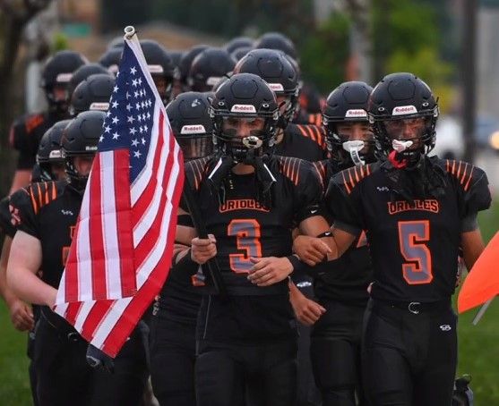 Young football players in black and orange uniforms march together, showcasing patriotism and team spirit before a game.