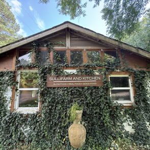 Charming rustic building adorned with ivy, wooden structure, large windows, sign "Sulfur Farm and Kitchen," serene atmosphere.