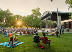 Outdoor concert in a park at sunset, with a joyful crowd enjoying music on the grass. A festive, communal atmosphere.