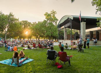 Outdoor concert in a park at sunset, with a joyful crowd enjoying music on the grass. A festive, communal atmosphere.