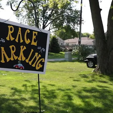 Yard sign reads "RACE PARKING" in bright yellow, with green grass and houses in the background, creating a fun, inviting vibe.