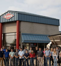 Group of diverse individuals in business attire stand outside a "KUHN" building, conveying teamwork and pride in agriculture.