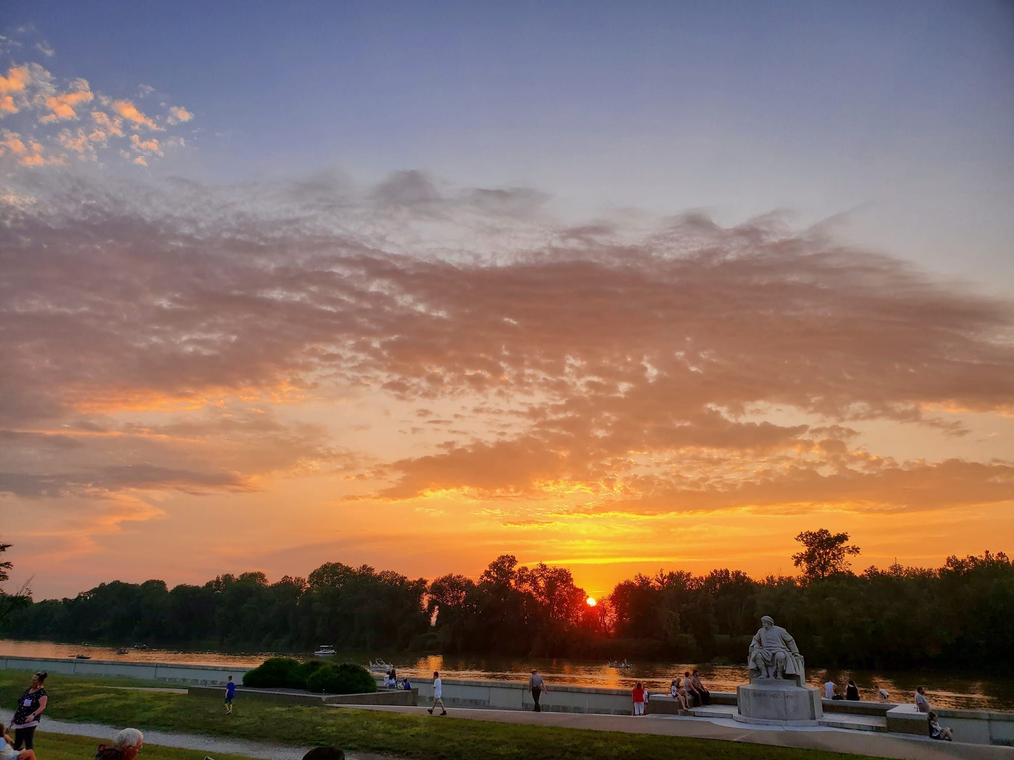 A serene sunset over water, with warm hues, scattered clouds, people enjoying the view, and lush greenery creating tranquility.