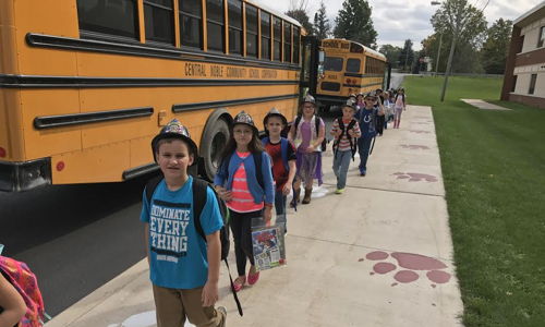 Children in colorful clothing walk happily in line on a sidewalk near a school bus, capturing a joyful school day moment.