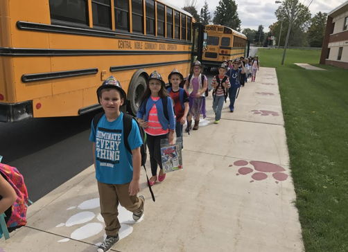 Children in colorful clothing walk happily in line on a sidewalk near a school bus, capturing a joyful school day moment.