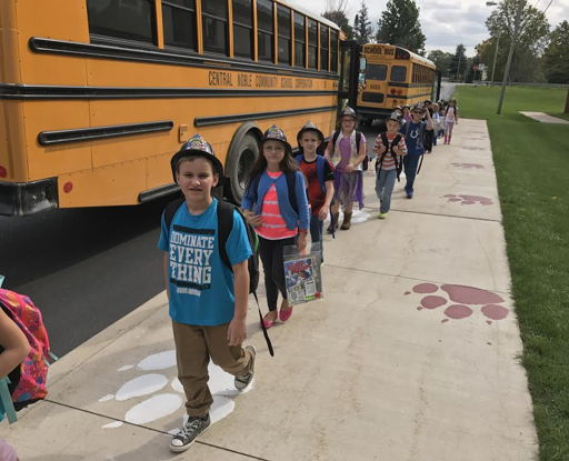 Children in colorful clothing walk happily in line on a sidewalk near a school bus, capturing a joyful school day moment.
