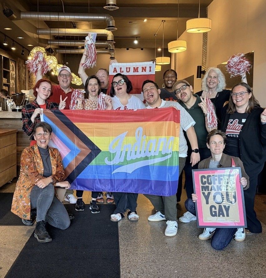 A joyful group in a cafe celebrates with pom-poms, signs, and a pride flag, embodying community and inclusivity.