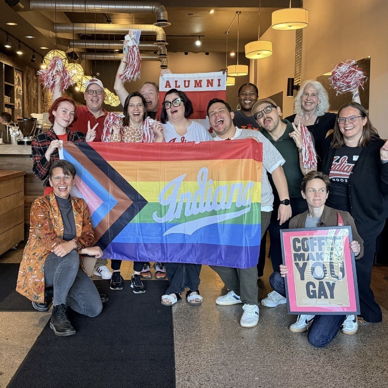 A joyful group in a cafe celebrates with pom-poms, signs, and a pride flag, embodying community and inclusivity.