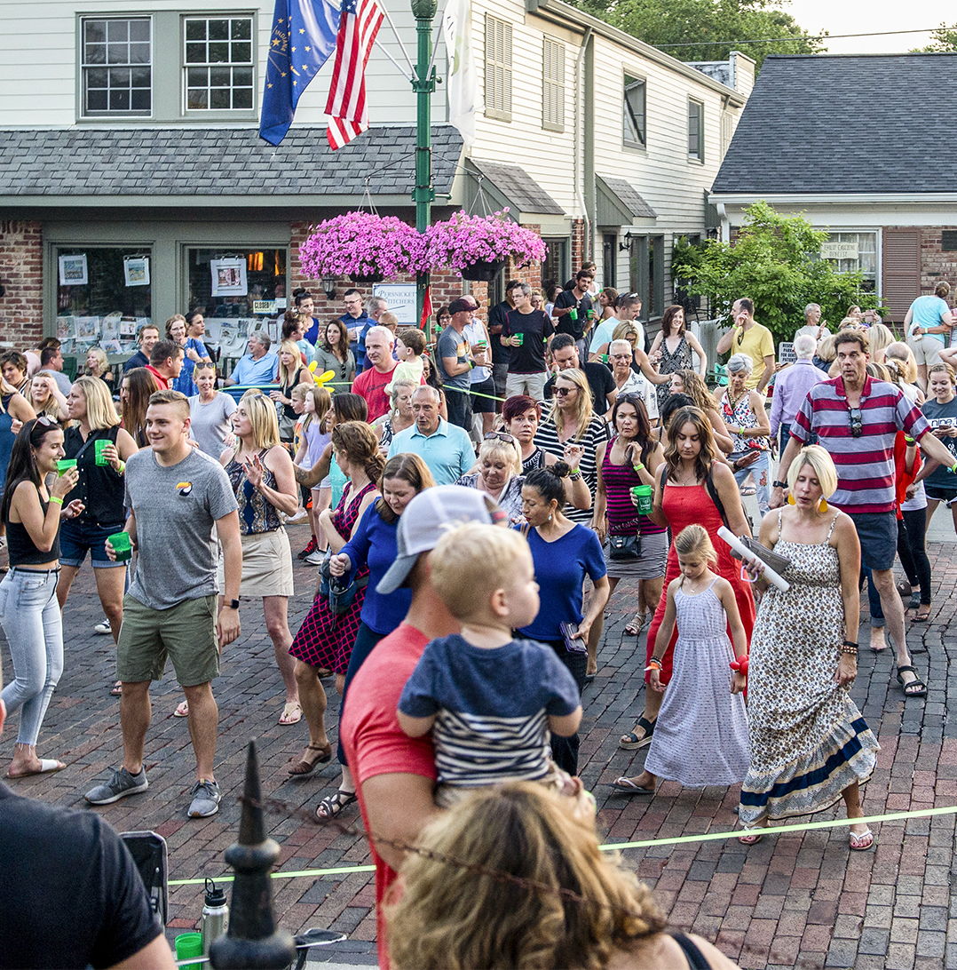 Outdoor gathering in a town square, with a diverse crowd celebrating joyfully, surrounded by flowers and American flags.