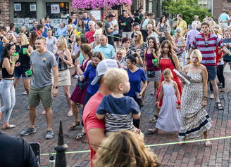 Outdoor gathering in a town square, with a diverse crowd celebrating joyfully, surrounded by flowers and American flags.