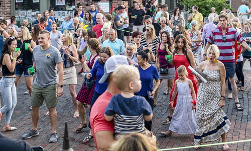 Outdoor gathering in a town square, with a diverse crowd celebrating joyfully, surrounded by flowers and American flags.