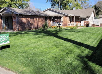 A vibrant front yard with a lush lawn, flower beds, and a “YARD OF THE MONTH” sign, set against sunny houses.