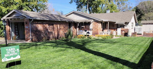 A vibrant front yard with a lush lawn, flower beds, and a “YARD OF THE MONTH” sign, set against sunny houses.
