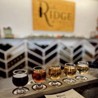 A flight of five colorful drinks on a wooden board, with a menu and "Ridge" sign, evokes warmth and social enjoyment.
