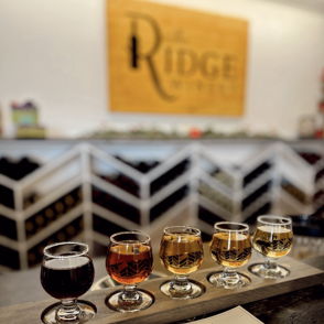 A flight of five colorful drinks on a wooden board, with a menu and "Ridge" sign, evokes warmth and social enjoyment.