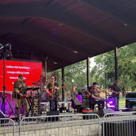 A vibrant outdoor live music performance with a band, crowd, large screen, American flag, and festive atmosphere.