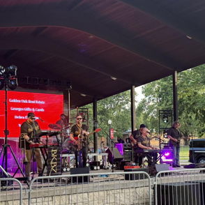 A vibrant outdoor live music performance with a band, crowd, large screen, American flag, and festive atmosphere.