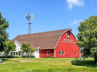 Charming rural scene with a red barn, white building, windmill, and serene pond under a blue sky, evoking tranquility.