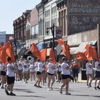 A lively parade with people in white and black holding orange flags in a vibrant downtown, capturing community spirit and joy.