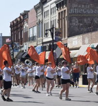 A lively parade with people in white and black holding orange flags in a vibrant downtown, capturing community spirit and joy.
