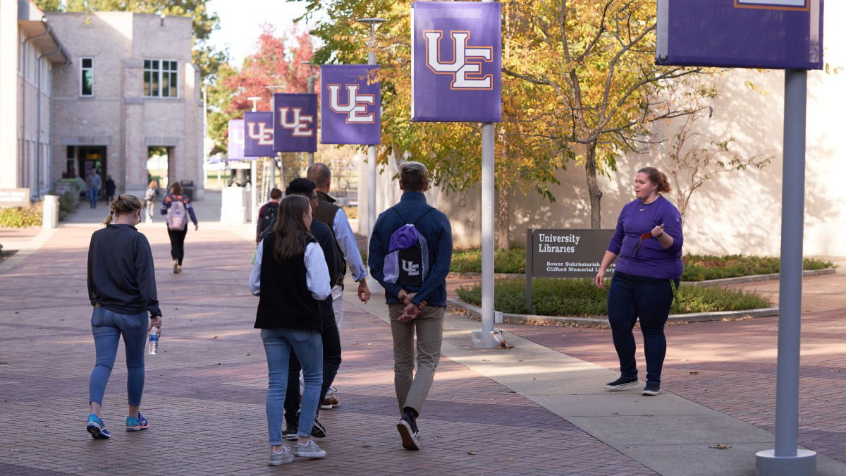 Campus scene with students walking, banners reading "UC," modern buildings, autumn trees, and a lively, inviting atmosphere.