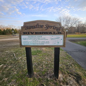 Wooden sign "Paradise Spring Riverwalk" in a serene natural landscape, suggesting a peaceful walking trail near water.