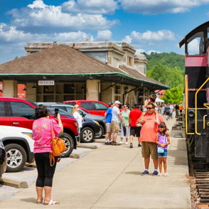 A vibrant pink train at a lively station, surrounded by people enjoying a cheerful, nostalgic atmosphere under a blue sky.