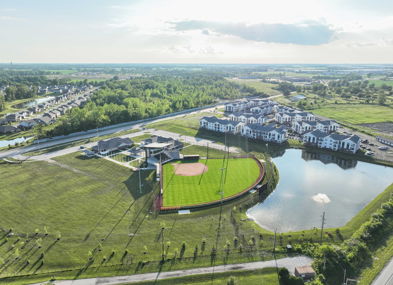 Aerial view of a serene suburban landscape with a baseball field, homes, a pond, and lush greenery, evoking tranquility and community.