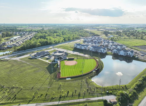 Aerial view of a serene suburban landscape with a baseball field, homes, a pond, and lush greenery, evoking tranquility and community.