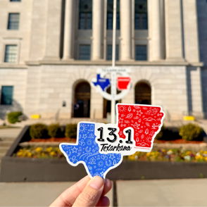 A hand holds a blue and red sticker of Texas and Arkansas in front of a classic building, evoking unity and patriotism.