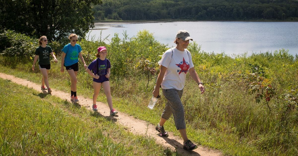 Four people walk along a sunny path by a serene lake, enjoying nature and each other's company in a cheerful atmosphere.