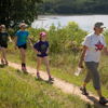 Four people walk along a sunny path by a serene lake, enjoying nature and each other's company in a cheerful atmosphere.