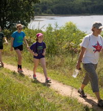 Four people walk along a sunny path by a serene lake, enjoying nature and each other's company in a cheerful atmosphere.